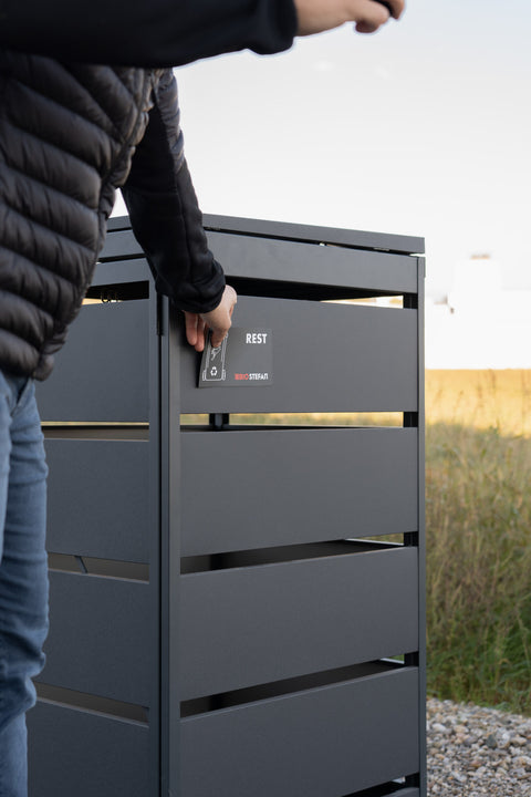 Person öffnet Bio Stefan Mülltonnenbox mit Karte, Logo sichtbar.
