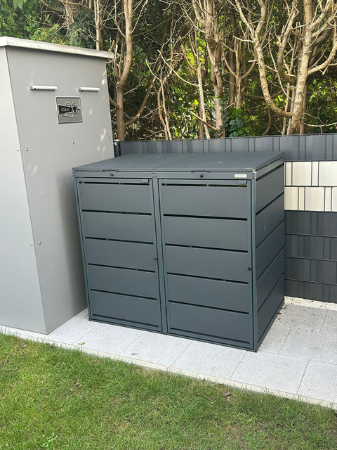 Two gray garbage can boxes from Bio Stefan stand on a paved area in front of trees.