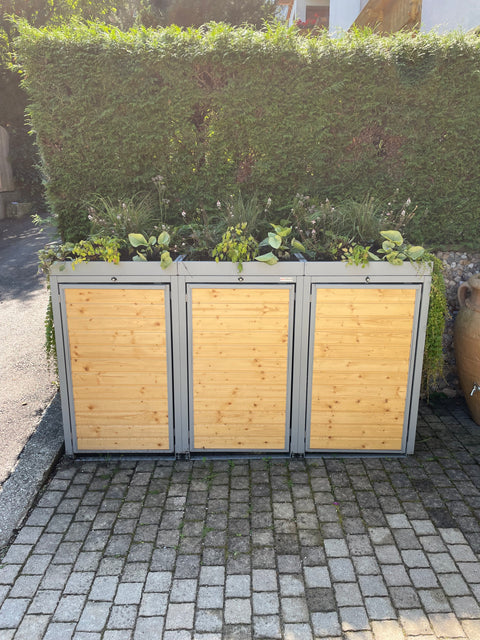 Mülltonnenbox aus Holz und Metall mit Pflanzdach von BIO Stefan vor grüner Hecke.