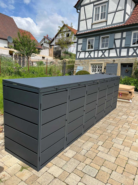 BIO Stefan Dagobert Mülltonnenbox 6er 240l in Anthrazit freistehend auf Pflaster im Hof vor einem Fachwerkhaus, frisch aufgebaut, daneben die Flatpack-Kartons