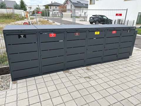BIO Stefan Dagobert Mülltonnenbox 6er 240l in Anthrazit frontal mit sechs beschrifteten Türen für Rest, Papier, Bio, Gelb, Papier und Rest vor Bauzaun und Neubauten