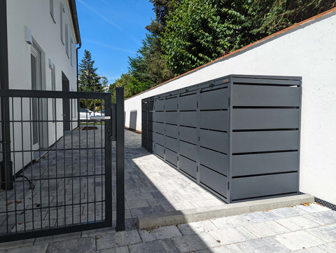 BIO Stefan Dagobert Mülltonnenbox 6er 240l in Anthrazit entlang einer weißen Hofmauer im gepflasterten Innenhof, Blick durch das geöffnete Gartentor