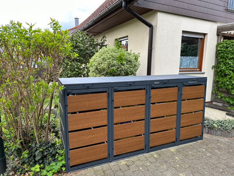 BIO Stefan Dagobert Mülltonnenbox 4er in Anthrazit mit Holzfront, freistehend vor Hausfassade zwischen Büschen