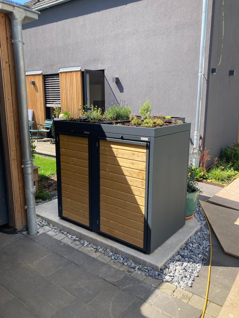 BIO Stefan Holzmichl Mülltonnenbox 2er 120 & 240 l in Anthrazit an der grauen Hauswand neben Regenrohr und Carport mit Pflanzdach