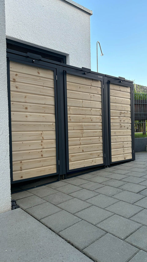 BIO Stefan Holzmichl Mülltonnenbox 3er 120l und 240l in Anthrazit mit Fichtenholz-Front entlang der hellen Hausfassade auf grauem Betonpflaster