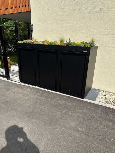 BIO Stefan Stahlfred Mülltonnenbox 3er 240l in Schwarz RAL 9005 mit bepflanztem Pflanzdach an der Hauswand neben schwarzem Doppelstabmattenzaun